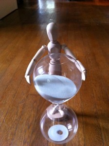 An hourglass with a mannequin on a hard wood floor.